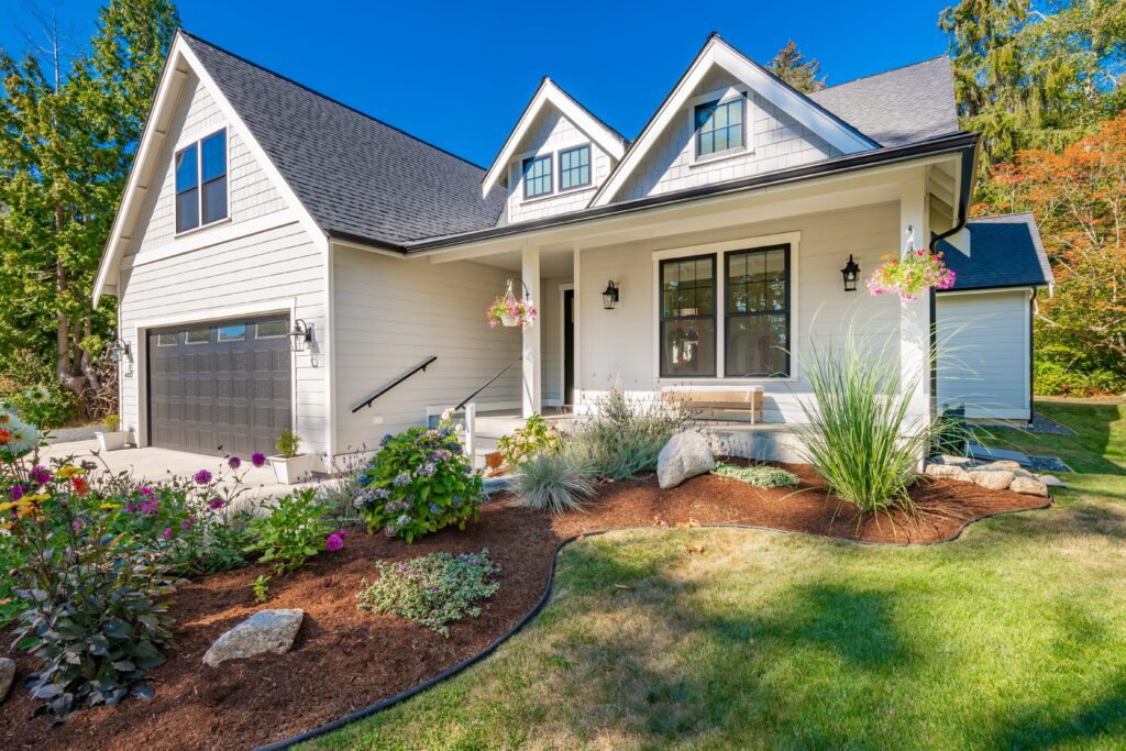 A white craftsman-style home with a dark asphalt shingle roof surrounded by colorful spring landscaping, illustrating the importance of residential roof maintenance in Kentucky.