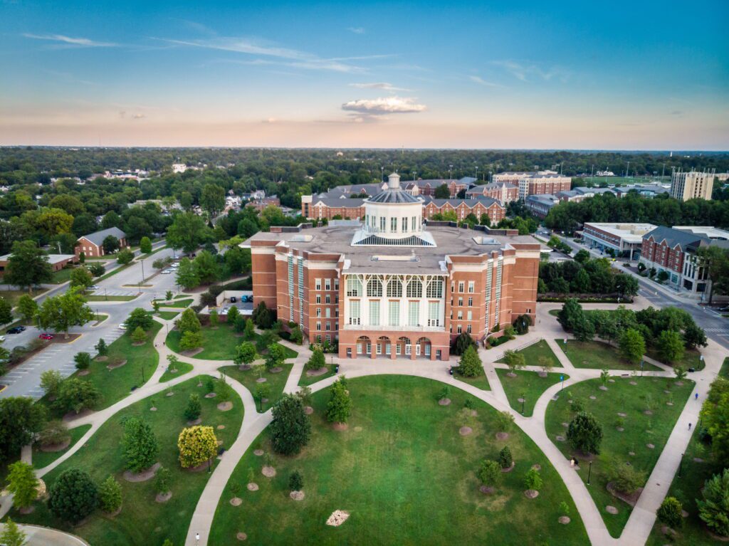 erial drone view of a large red-brick university library building in lexington, ky with a distinctive glass rotunda on its roof, surrounded by manicured green lawns, curved walkways, and scattered trees on a college campus, with additional brick academic buildings and a wooded suburban landscape visible in the background under a partly cloudy blue sky.